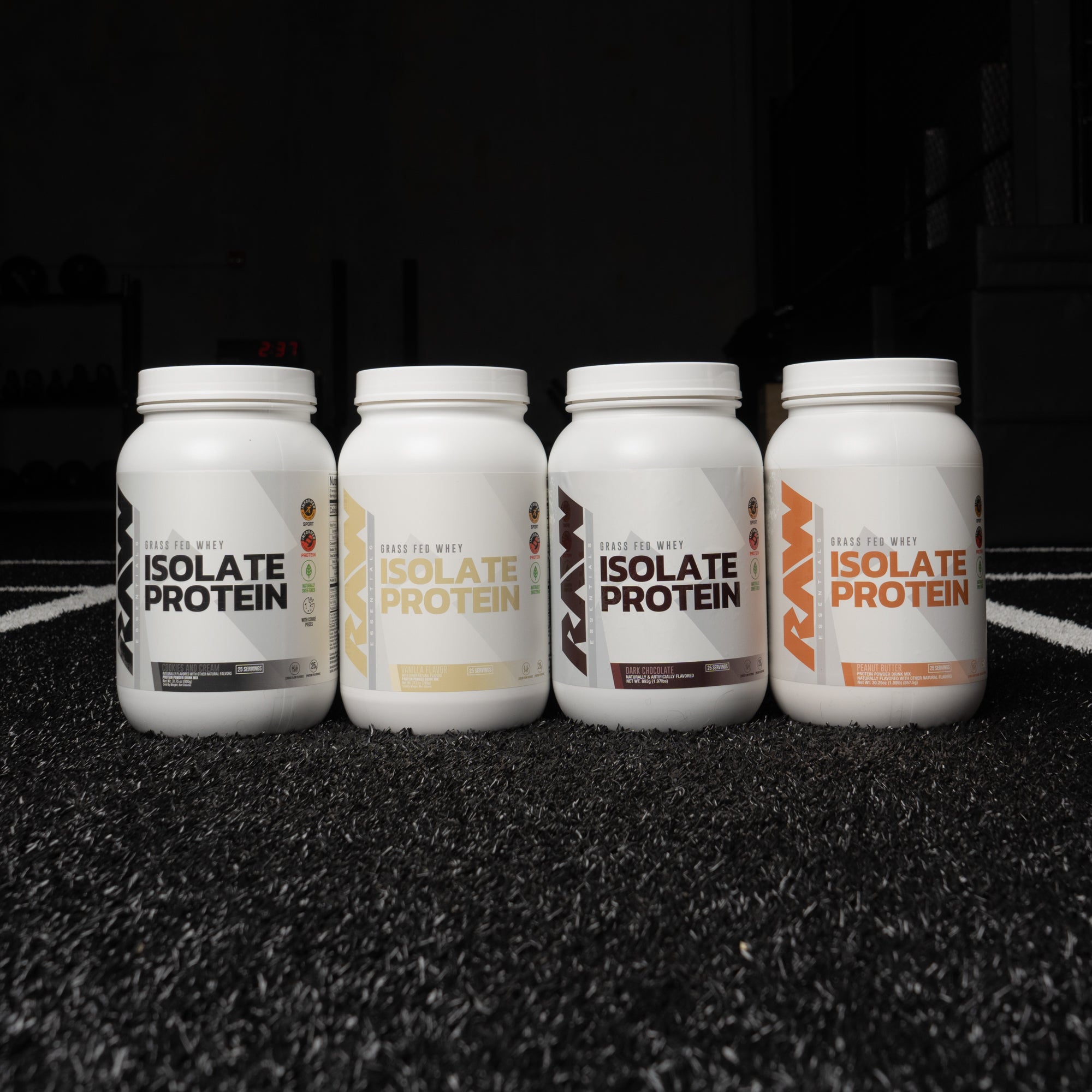 Four RAW Isolate Protein containers in different flavors are displayed side by side on a textured black gym floor.