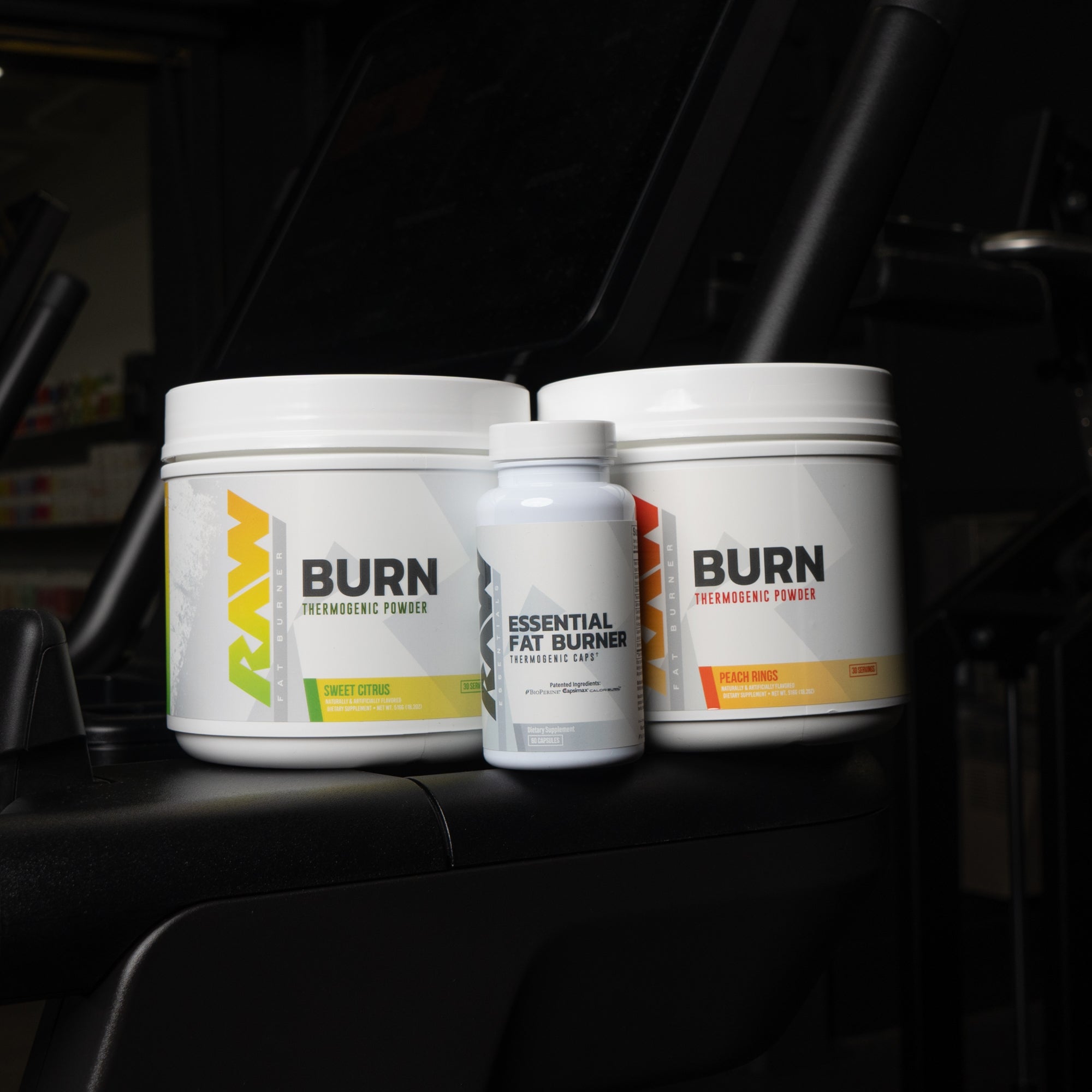 RAW Nutrition's Burn thermogenic powders in Sweet Citrus and Peach Rings flavors, along with Essential Fat Burner capsules, displayed on a black exercise machine in a gym environment.