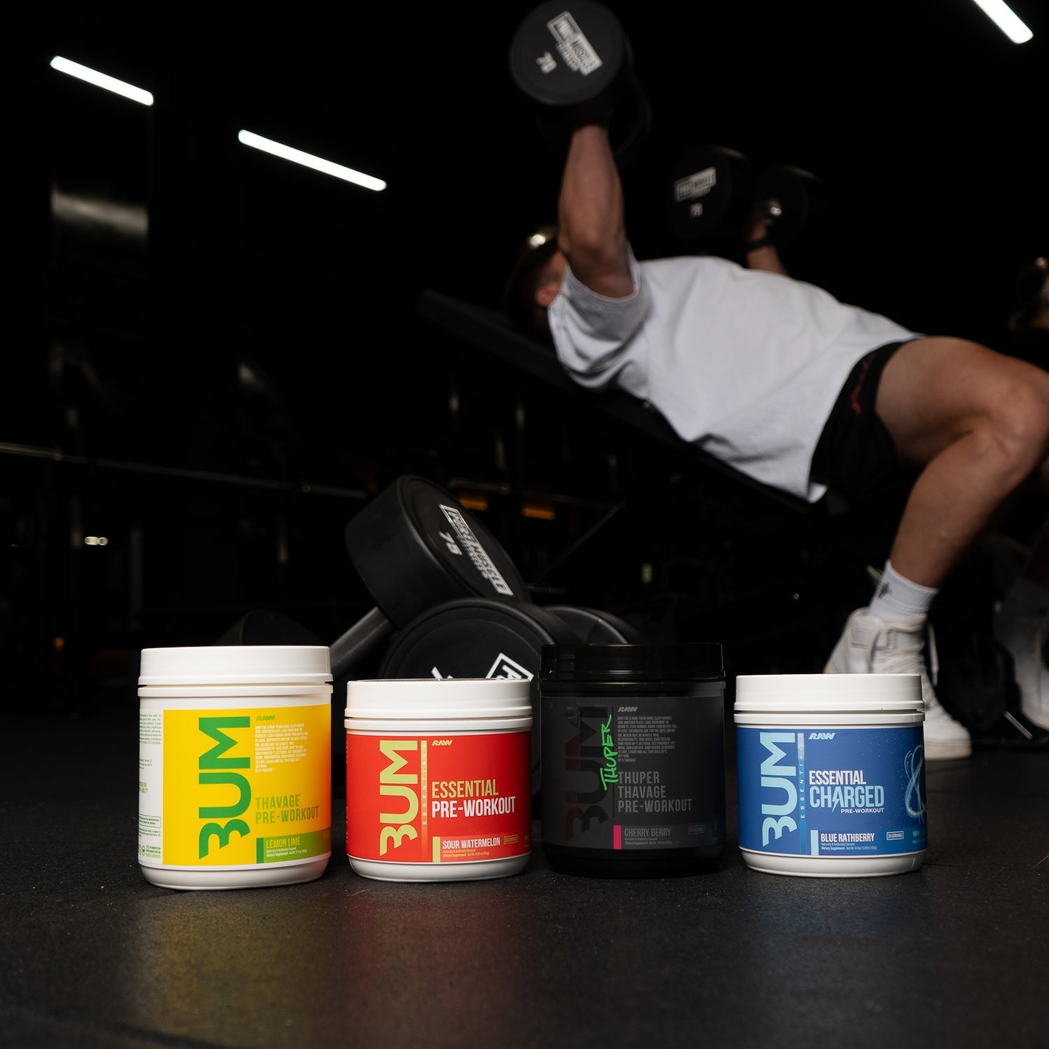 Four RAW pre-workout products, including Thavage, Essential Pre-Workout, Thuper Thavage, and Essential Charged, are displayed on a gym floor with a person performing a dumbbell press in the background.