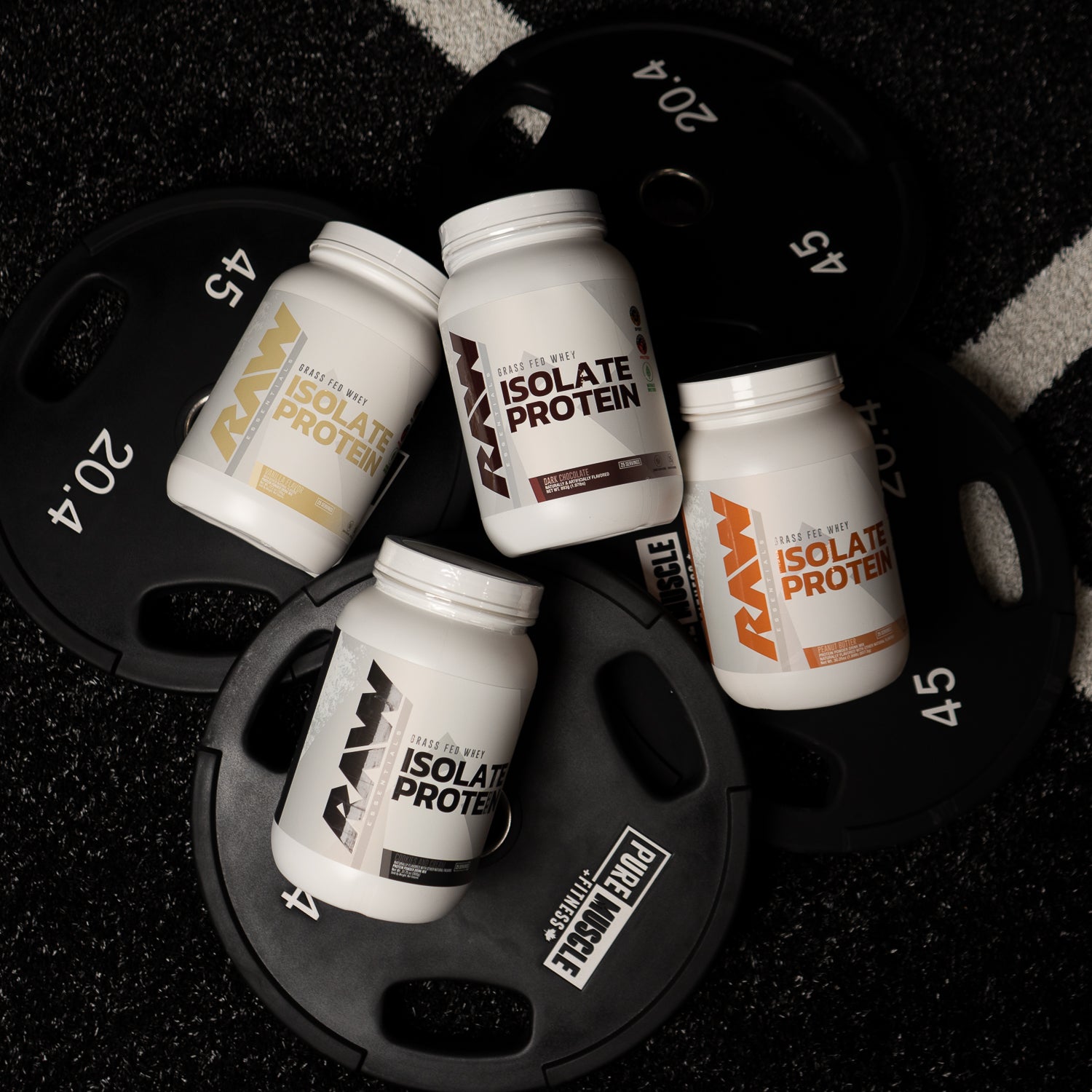 Four containers of RAW Isolate Protein in different flavors are arranged on black weight plates, set against a textured gym floor.