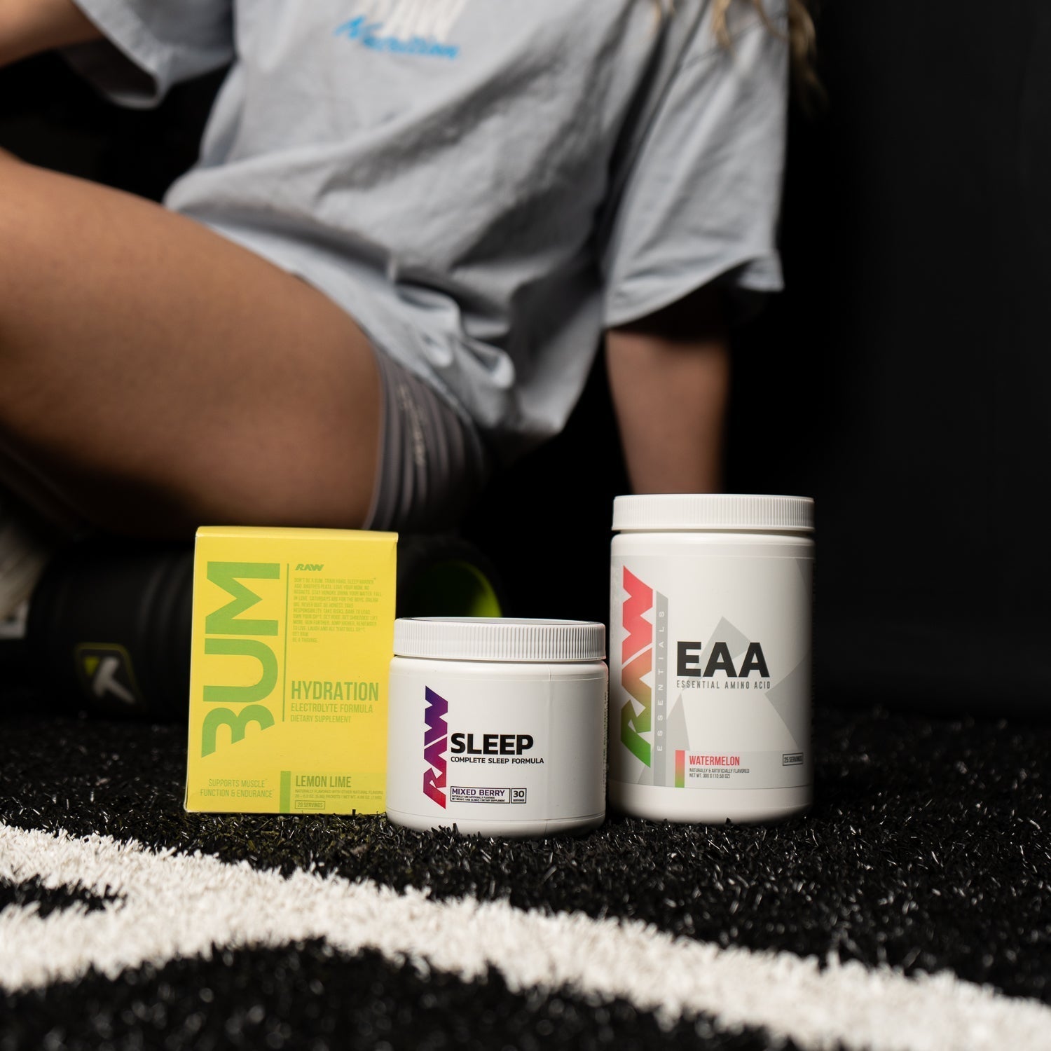 RAW Hydration, Sleep, and EAA supplements displayed on a turf gym floor with an athlete sitting in the background.