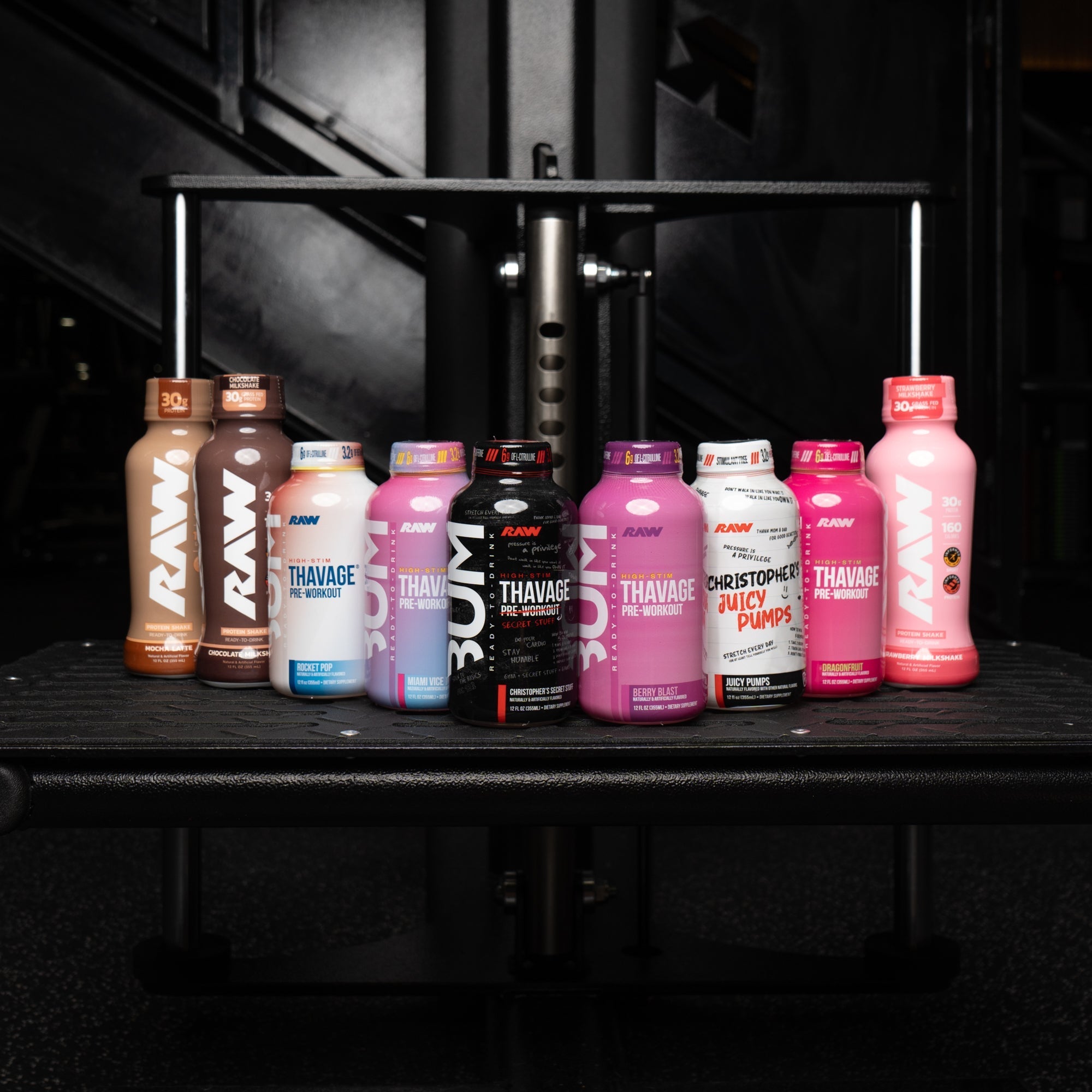 An assortment of RAW Nutrition ready-to-drink products, including protein shakes and pre-workouts in various flavors, are displayed on a black gym bench, set against the backdrop of gym equipment.