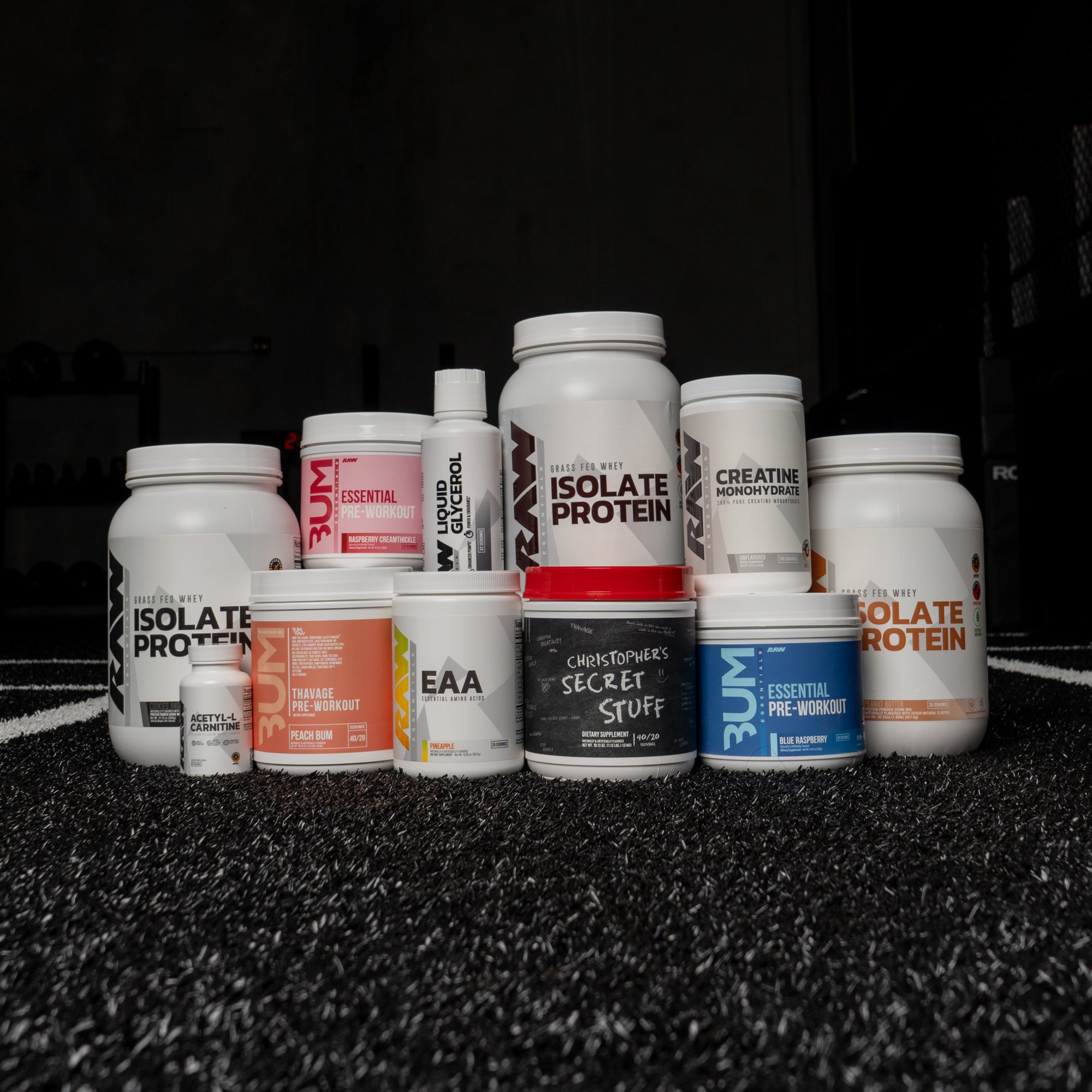 A selection of RAW Nutrition supplements, featuring Isolate Protein, Essential Pre-Workout, Creatine Monohydrate, Liquid Glycerol, and more, neatly arranged on a textured black gym floor.