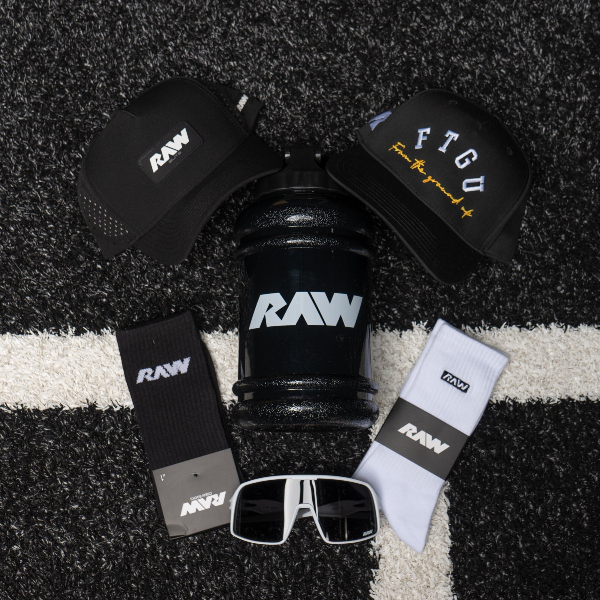 A collection of RAW accessories displayed on a turf background, featuring branded caps, a large black water jug, black and white crew socks, and sunglasses.
