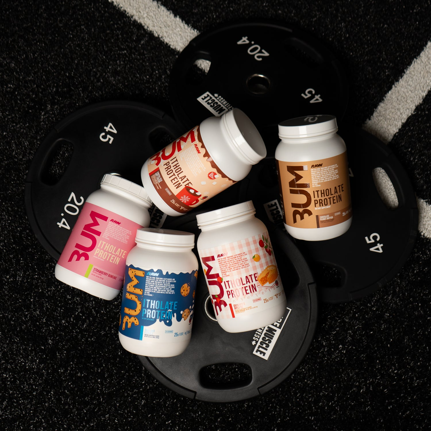 An assortment of RAW Itholate Protein containers in various flavors is creatively arranged on black weight plates, set against a textured gym floor.