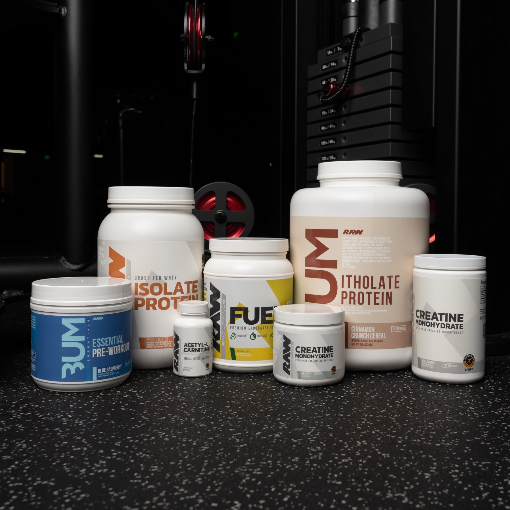 A collection of RAW Nutrition supplements, including Isolate Protein, Itholate Protein, Essential Pre-Workout, Fuel, Creatine Monohydrate, and Acetyl-L-Carnitine, displayed on a gym floor with weight equipment in the background.
