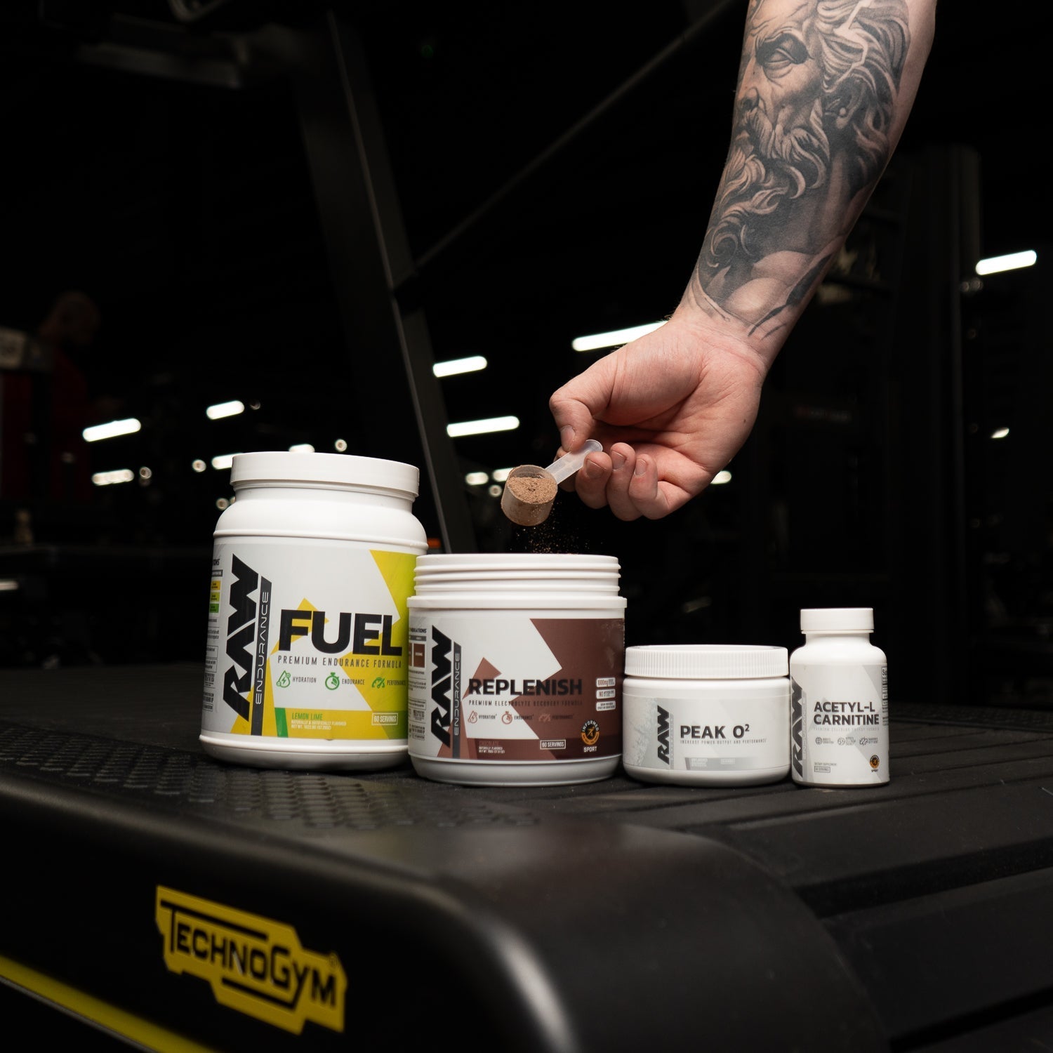 A tattooed arm scoops powder into a RAW Replenish container, with additional RAW supplements including Fuel, Peak O2, and Acetyl-L-Carnitine displayed on a gym treadmill under low lighting.