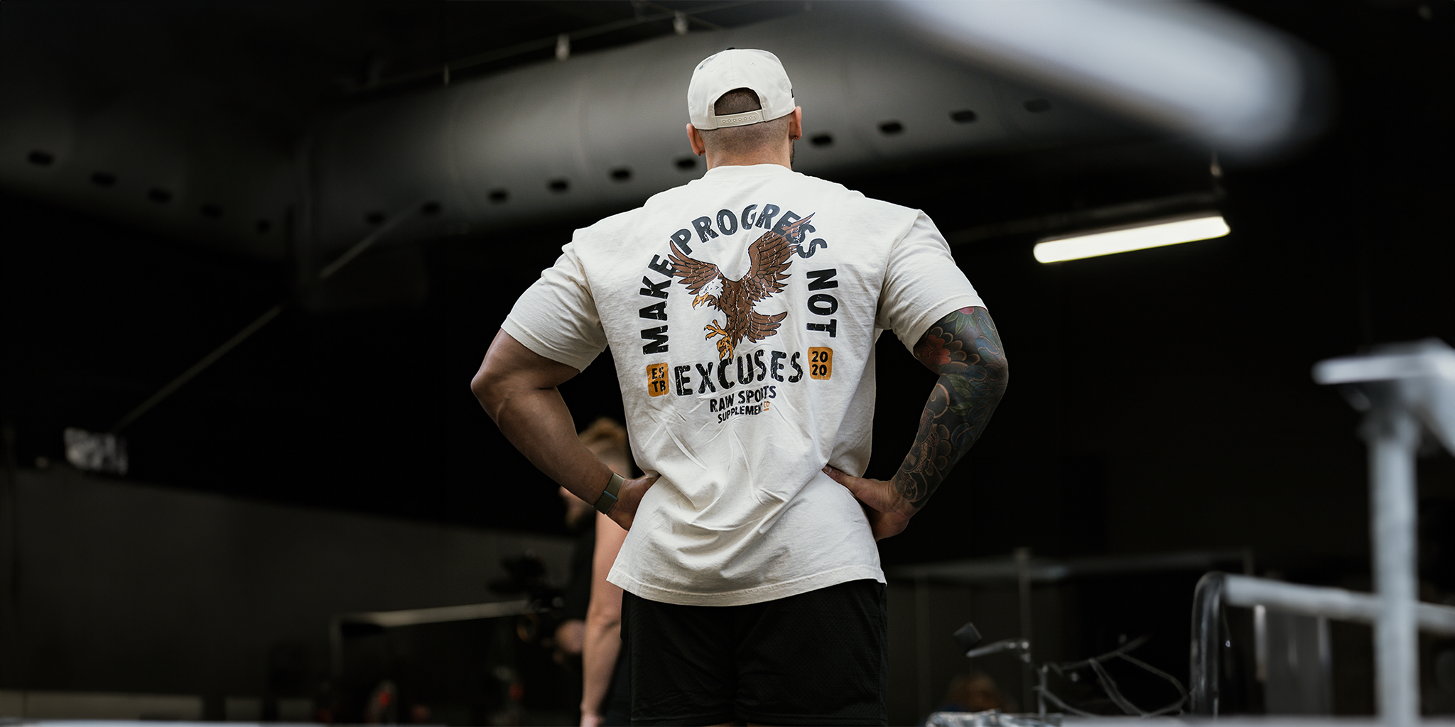 A muscular man in a gym wearing a white T shirt with the slogan Make Progress Not Excuses featuring an eagle graphic and the RAW Sports Supplements logo viewed from behind under dark lighting.