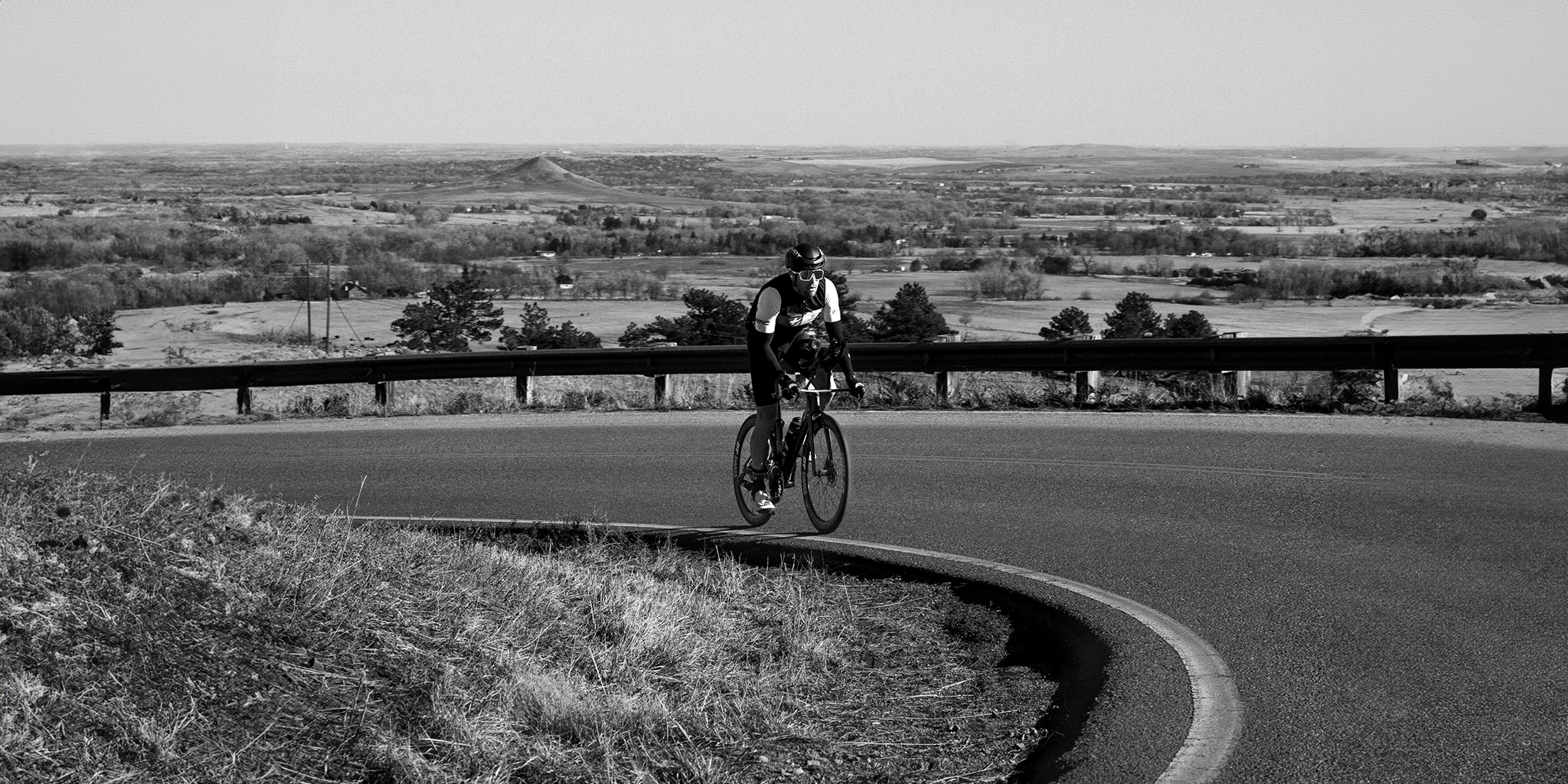A male cyclist wearing a helmet and aerodynamic gear riding uphill on a winding rural road with a wide landscape of fields and hills in the background. The image conveys themes of endurance, focus, and outdoor athletic performance.
